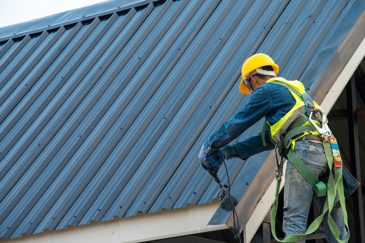 Construction worker wearing safety harness belt using air or pneumatic nail gun and installing metal roof tile on top new roof.